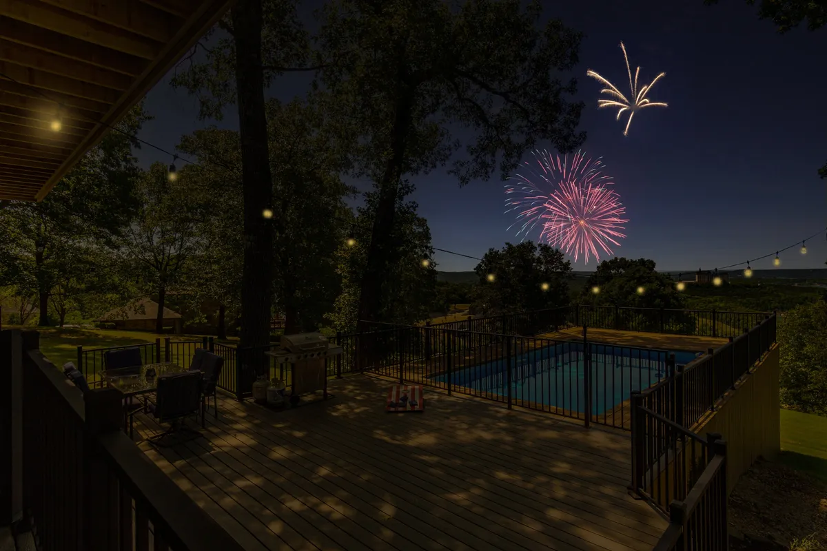 A vibrant exterior shot showing fireworks lighting up the night sky above a swimming pool, with string lights and inviting deck furniture creating a lively and festive atmosphere.