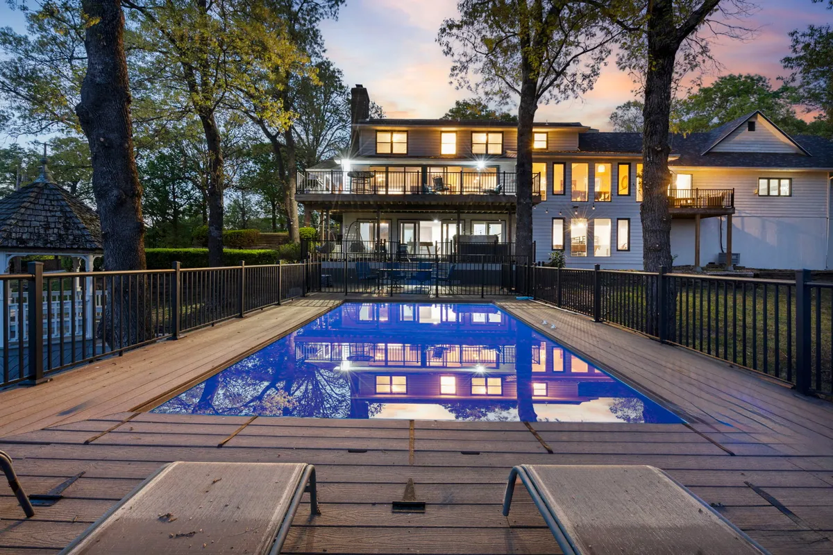 The image showcases a stunning backyard featuring a sparkling outdoor saltwater pool with two inviting lounge chairs, a spacious deck for entertaining, and the elegant facade of a large single-family home. The scene is set for relaxation and socializing, with a tranquil lake view backdrop and the warm hues of sunset reflected on the water.
