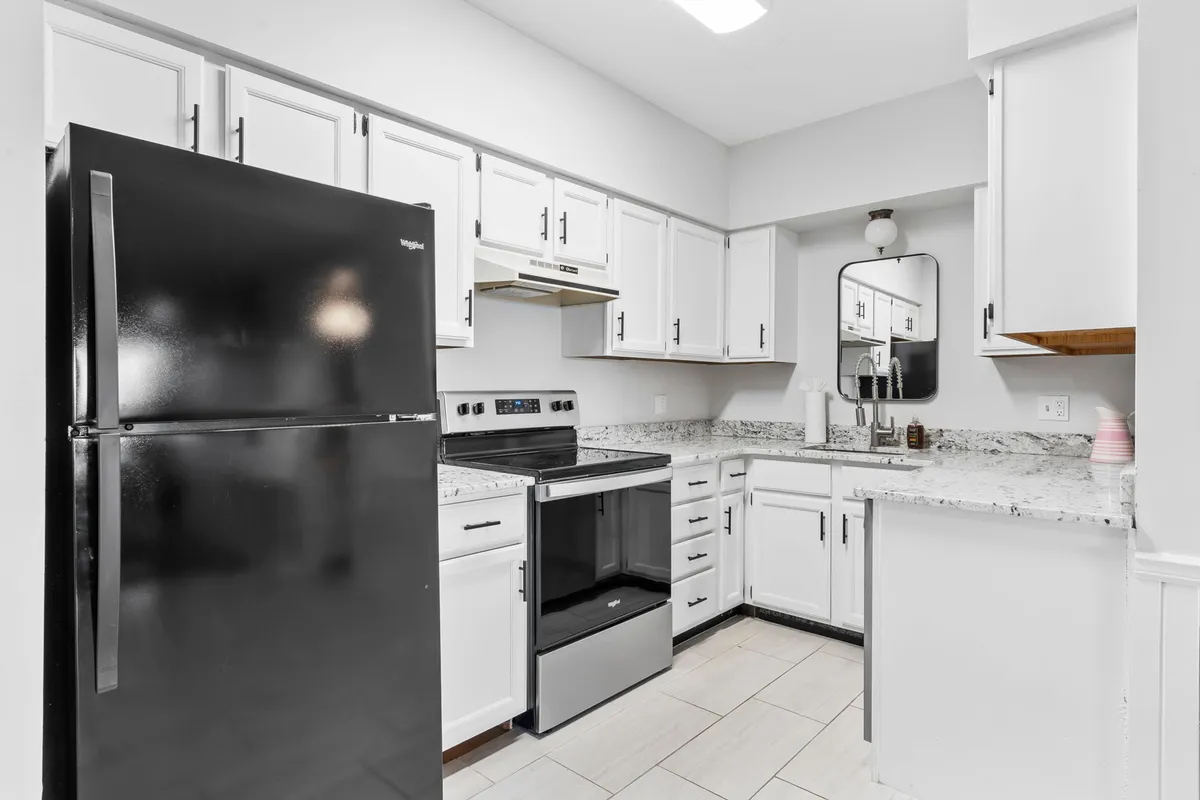 A spacious, modern kitchen with a black refrigerator, stainless steel stove, white cabinetry, and a granite countertop. The mirror above the sink adds depth, while the layout is functional and inviting for group cooking or meal prep.
