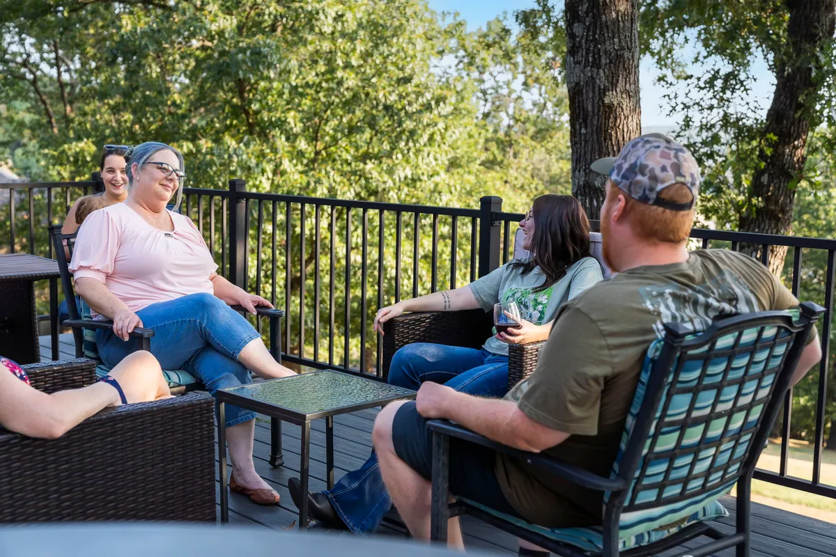 A lively group of friends is gathered in an outdoor seating area, surrounded by lush greenery and trees, sharing drinks and conversation in a relaxed, inviting setting. The scene captures the spirit of togetherness and leisure in nature.