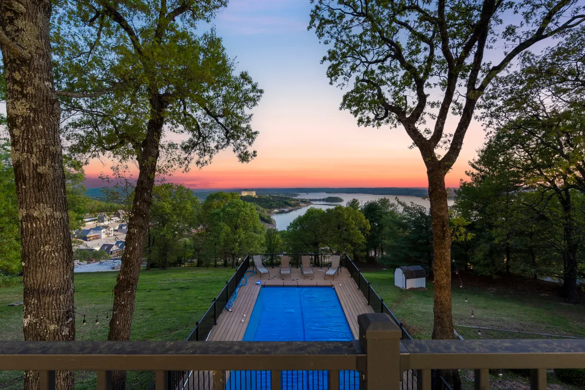 This exterior image showcases a serene swimming pool set against a backdrop of trees and a breathtaking lake view. The sunset sky casts a warm glow over the entire scene, while the poolside area looks inviting for both people and pets to relax and unwind after a day outside.