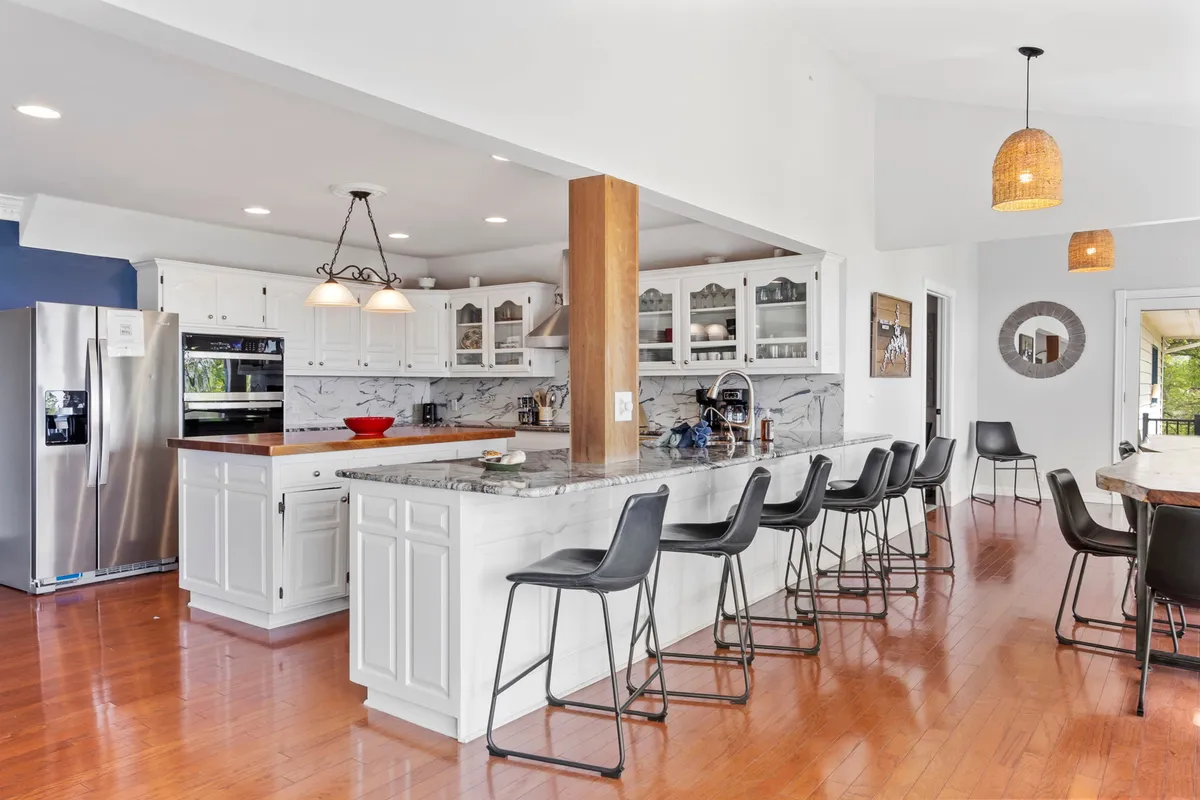 This image showcases a welcoming kitchen with a central island, stainless steel appliances, bar stools, and marble countertops. The space exudes an inviting and social vibe, perfect for cooking, entertaining guests, and gathering for meals.
