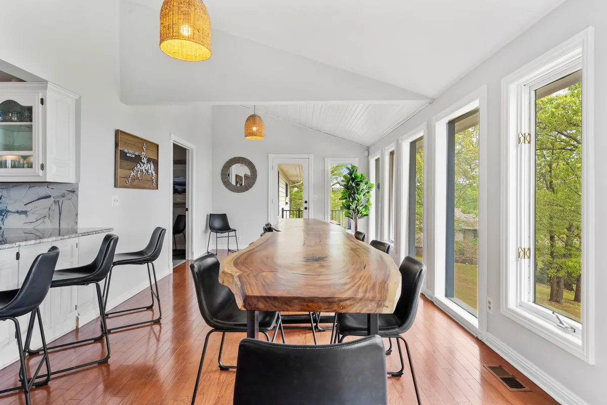 A large wooden dining table anchors a sunlit dining area, surrounded by windows that offer lush greenery views. Pendant lighting creates a warm, intimate setting for family meals or festive gatherings.