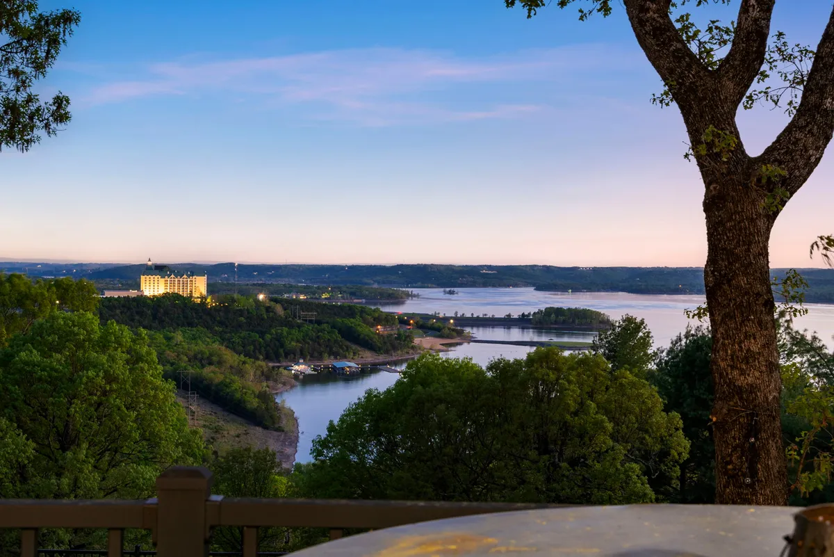 A wide view captures a river bordered by trees, a bridge, and a resort hotel, evoking a sense of peaceful adventure. The scene invites thoughts of boating, fishing, picnics, or simply watching the sunset over the water.