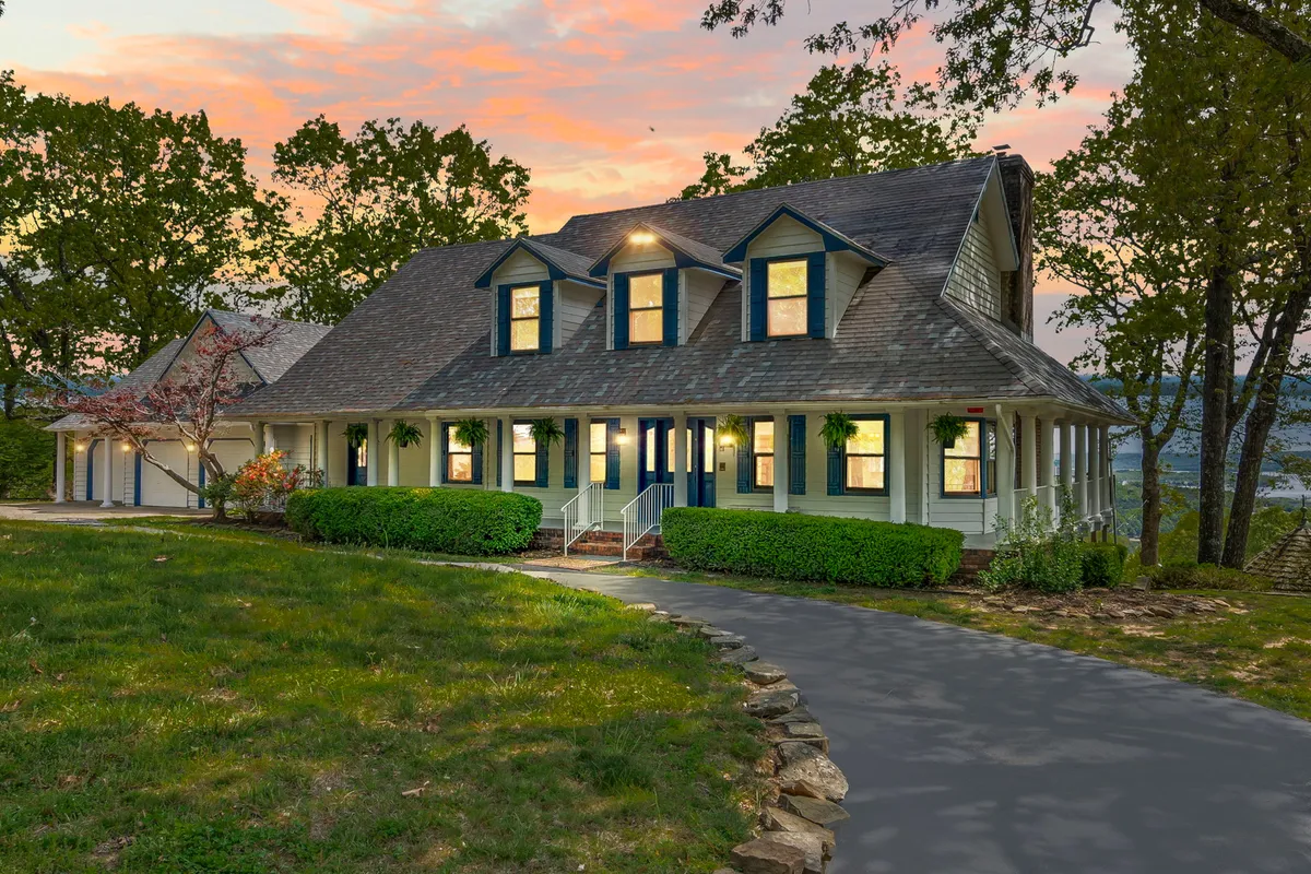 This image showcases the full façade of the home with its inviting wrap-around porch, large windows, and a pathway leading through mature trees. The setting feels peaceful and private, ideal for gatherings or contemplative evenings.