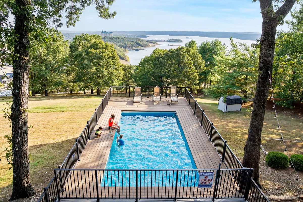 This exterior shot showcases a sparkling swimming pool with a person enjoying the water, surrounded by trees and featuring a stunning view of the lake in the background. There is ample space for lounging and socializing around the pool area, evoking a sense of relaxation and group leisure.