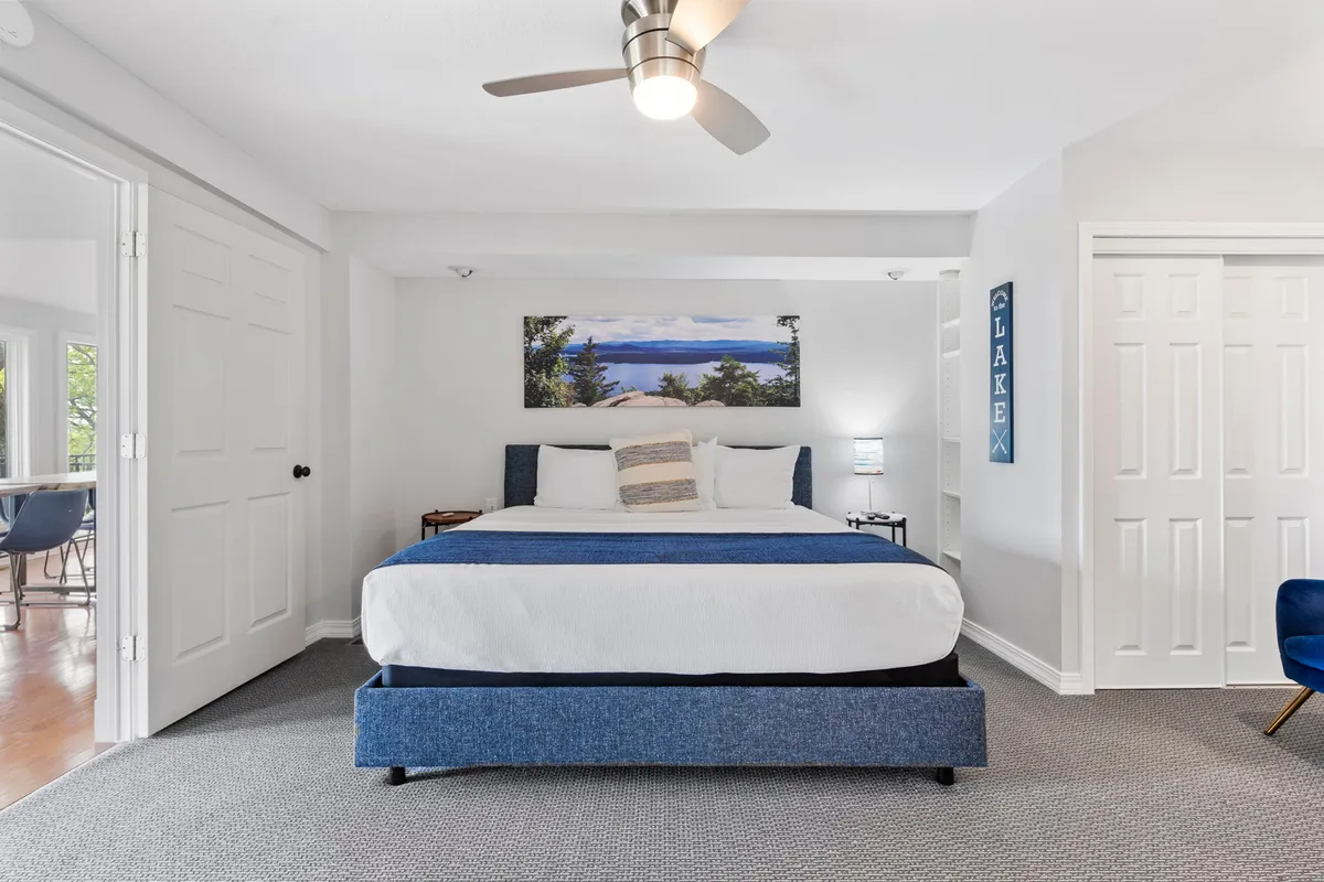 A serene bedroom featuring a large bed with a blue frame, soft bedding, a blue accent chair, and calming landscape artwork above the bed. Warm sunlight streams in, creating a peaceful, inviting atmosphere ideal for restful retreats.