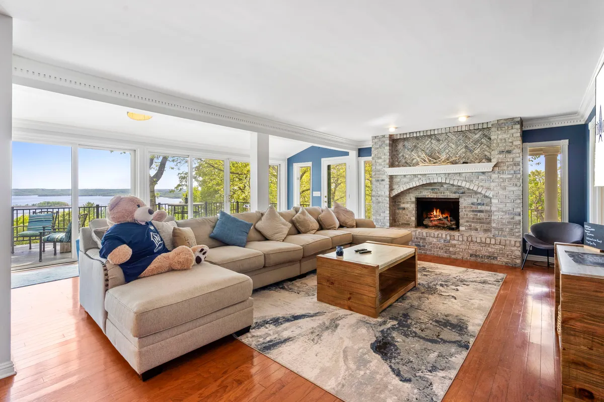 The family room features a plush sectional sofa, a stone fireplace, and expansive windows framing serene lake views. The setup is cozy and inviting, designed for socializing, relaxation, and enjoying the panoramic scenery.
