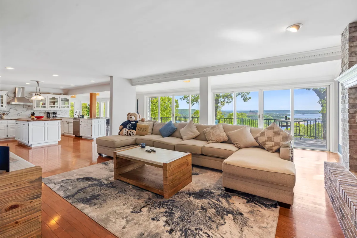 A large sectional sofa wraps around a wooden coffee table, with a teddy bear adding a playful touch. Large windows flood the space with light and offer a scenic view, creating a perfect spot for family gatherings or movie marathons.