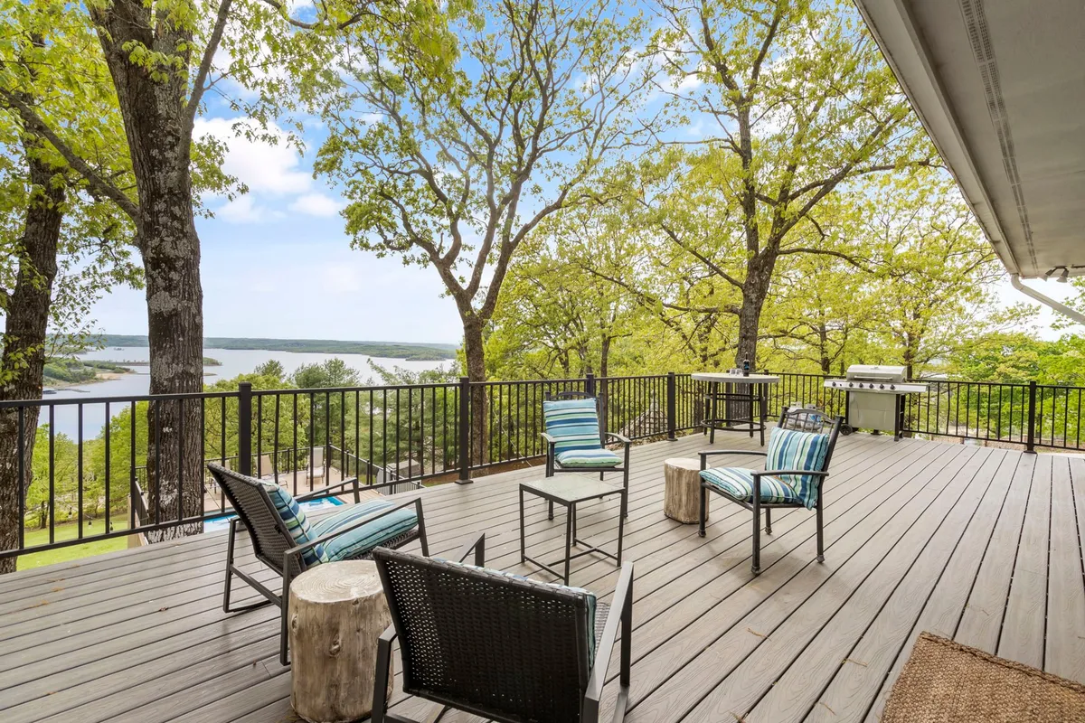 A wide wooden deck overlooks the lake, surrounded by trees and equipped with comfortable deck seating and a grill area. It invites guests to relax, enjoy outdoor meals, and take in the tranquil views with their pets at their side.
